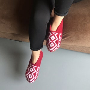 May include: Red and white knitted slippers with a geometric pattern. The slippers are worn on a person's feet.