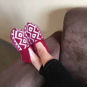 May include: Red and white knitted slippers with a geometric pattern. The slippers are worn by a person sitting on a brown couch.