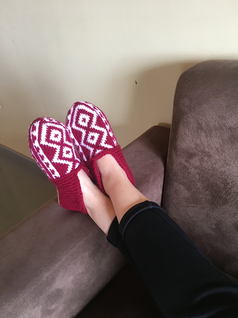 May include: Red and white knitted slippers with a geometric pattern. The slippers are worn by a person sitting on a brown couch.