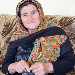 May include: An older woman wearing a colorful headscarf sits on a couch and knits with gray yarn.