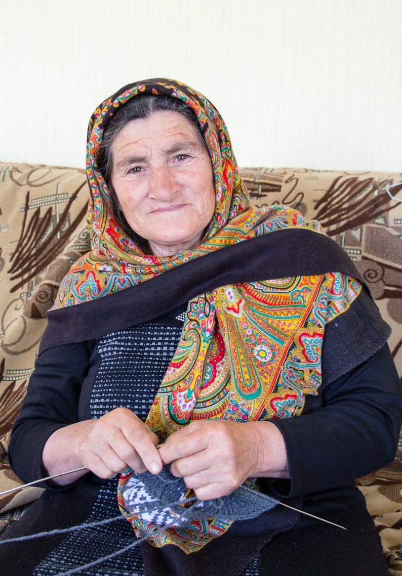 May include: An older woman wearing a colorful headscarf sits on a couch and knits with gray yarn.