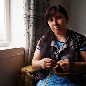 May include: A woman sits in a chair and knits with yellow yarn. She is wearing a brown and gray striped shirt and a blue shirt with a bird pattern underneath.