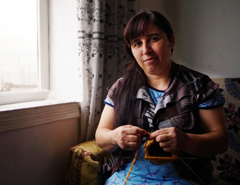 May include: A woman sits in a chair and knits with yellow yarn. She is wearing a brown and gray striped shirt and a blue shirt with a bird pattern underneath.