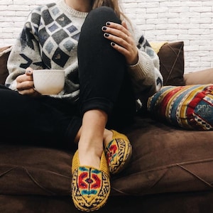 May include: A person wearing a white and black patterned sweater and black pants sits on a brown couch with a colorful pillow. They are holding a white mug and wearing yellow and blue patterned slippers.