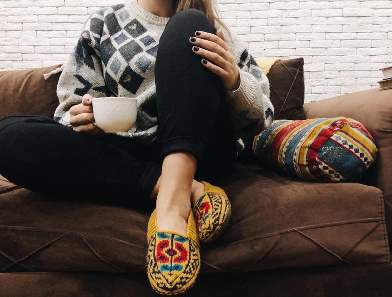 May include: A person wearing a white and black patterned sweater and black pants sits on a brown couch with a colorful pillow. They are holding a white mug and wearing yellow and blue patterned slippers.