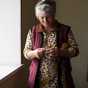 May include: A woman wearing a maroon vest over a brown and yellow patterned shirt is working on a craft project. She is holding a needle and thread and has several spools of colorful yarn in her hands.