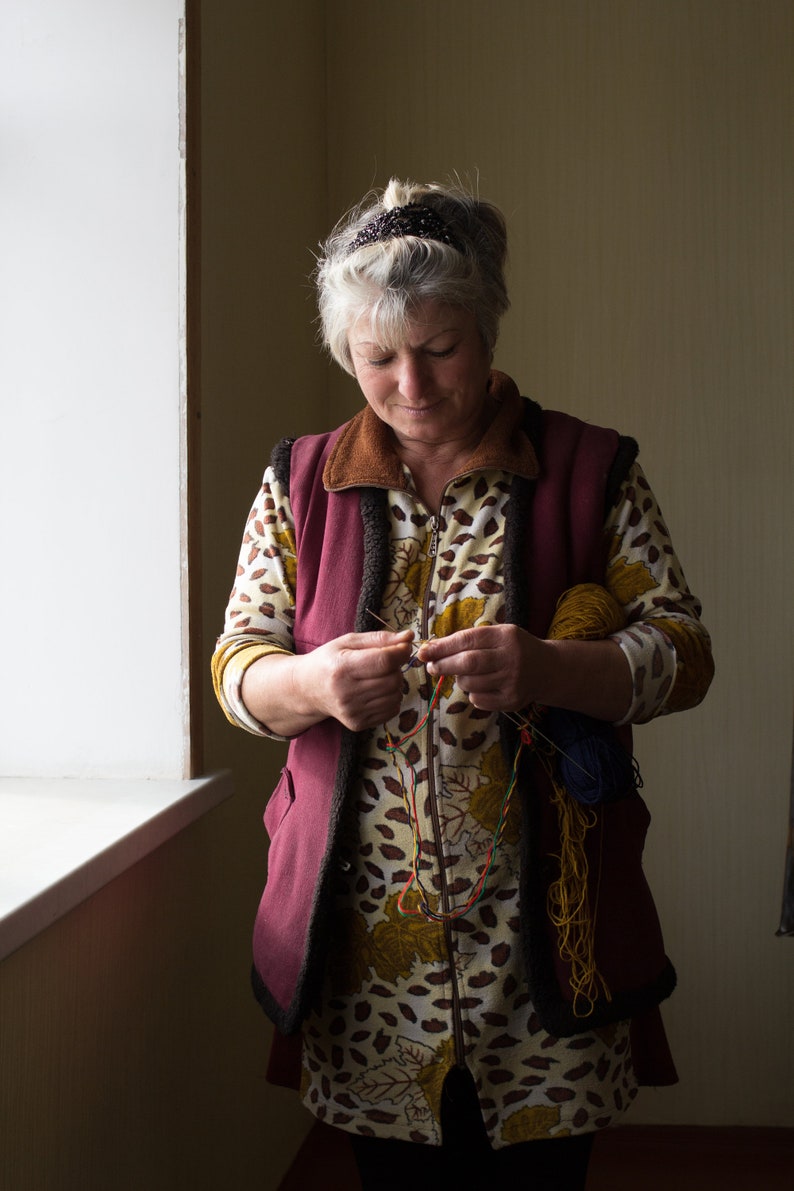 May include: A woman wearing a maroon vest over a brown and yellow patterned shirt is working on a craft project. She is holding a needle and thread and has several spools of colorful yarn in her hands.