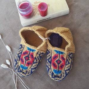 May include: A pair of knitted slippers in a mustard yellow color with a blue, red, and turquoise geometric pattern. The slippers are made of wool and have a comfortable fit.