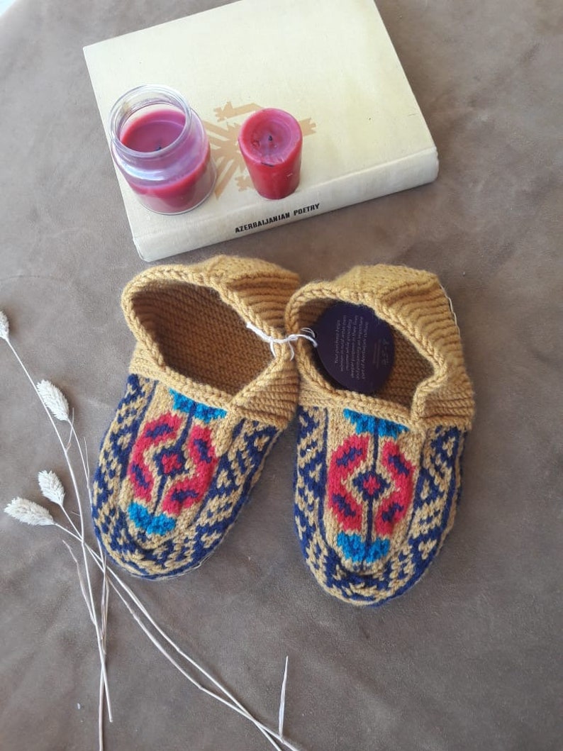May include: A pair of knitted slippers in a mustard yellow color with a blue, red, and turquoise geometric pattern. The slippers are made of wool and have a comfortable fit.