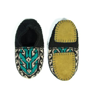 May include: A pair of black knitted slippers with a teal and beige geometric pattern. The slippers have a suede sole with a stitched edge.