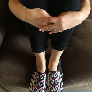May include: A pair of black and white knitted slippers with red accents. The slippers have a geometric pattern and are worn by a person sitting on a brown couch.
