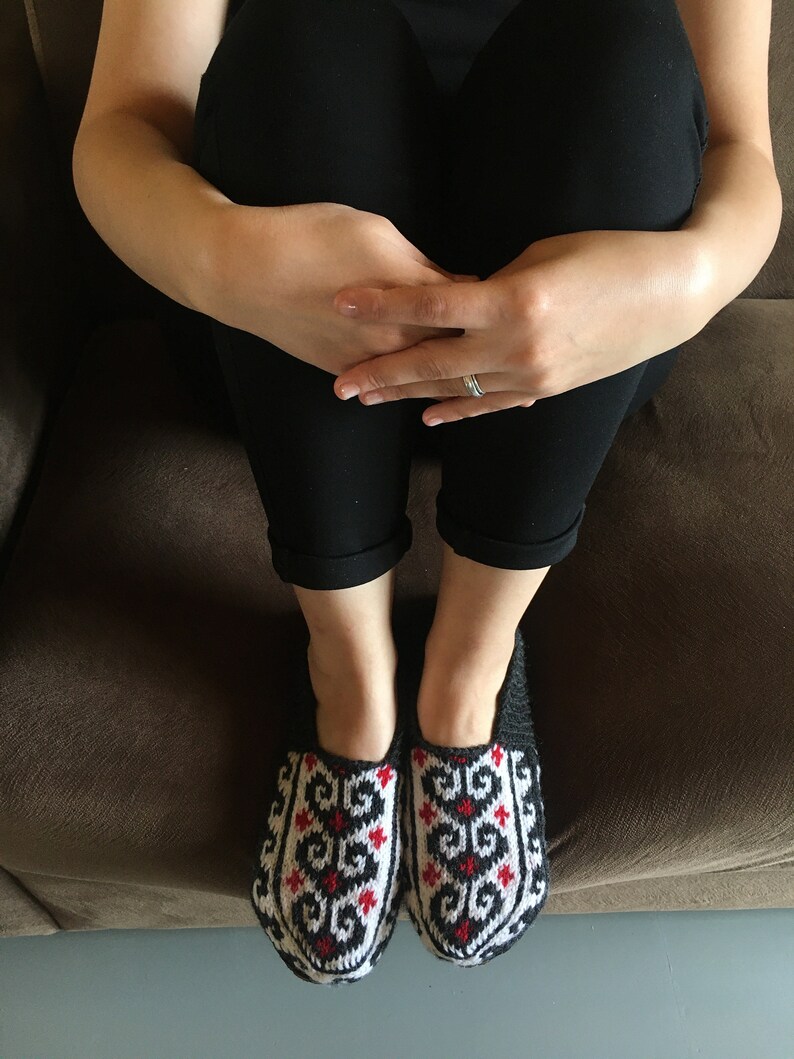 May include: A pair of black and white knitted slippers with red accents. The slippers have a geometric pattern and are worn by a person sitting on a brown couch.