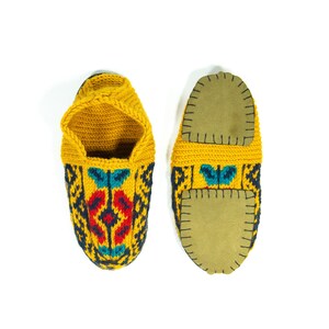 May include: A pair of yellow knitted slippers with colorful geometric patterns and brown leather soles.