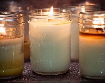 May include: A group of clear glass mason jars containing white wax candles. The center candle is lit with a visible flame. Each jar features the embossed Ball logo and the words Wide Mouth on the glass surface. The wax appears smooth and solid.