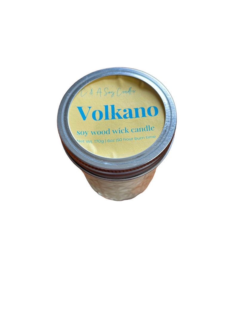 May include: A yellow and blue soy wood wick candle in a glass jar with a silver lid. The label reads "Volkano soy wood wick candle Net Wt. 170g | 6oz 150 hour burn time".