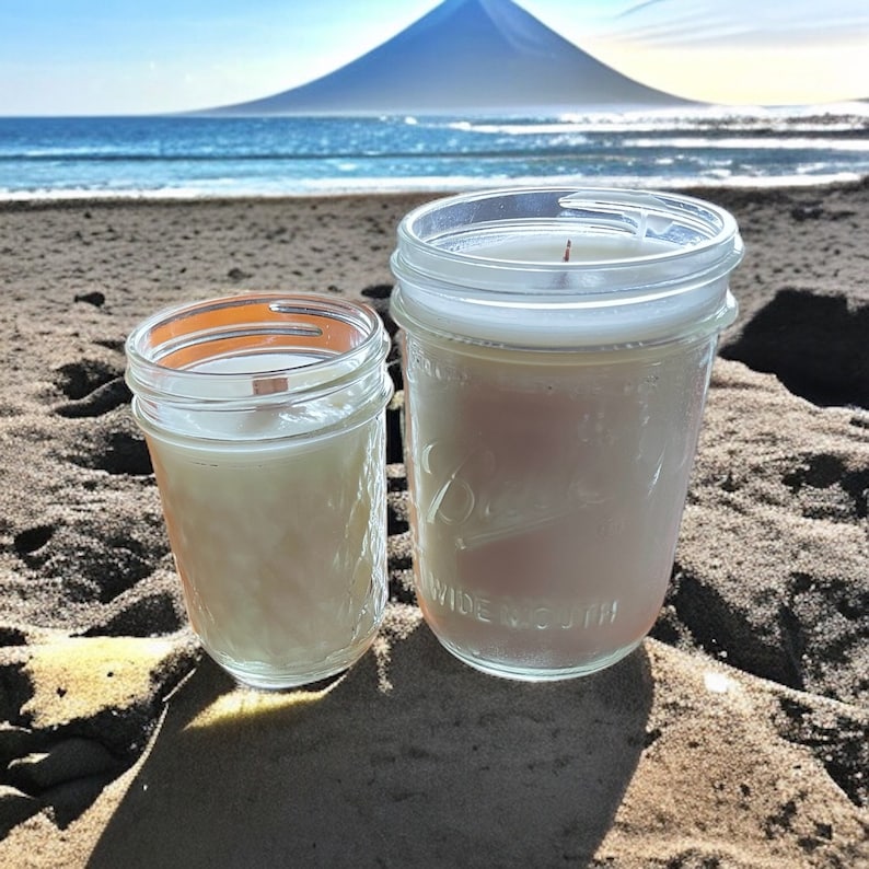 May include: Two glass jars containing white candles, positioned on a sandy beach with a mountain in the distance.