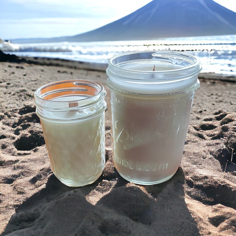 May include: Two glass jars containing white candles, resting on a sandy beach with a mountain in the distance.