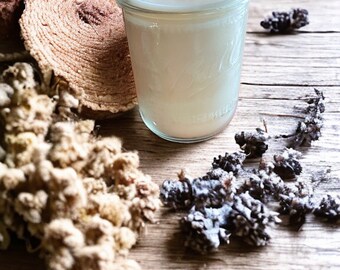May include: A white candle in a clear glass jar with a wooden wick. The candle is surrounded by dried flowers and other natural elements on a wooden surface.