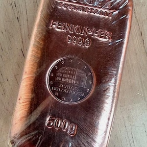 May include: A copper bar in plastic wrapping. The bar is stamped with "GEIGER EDELMETALLE", "FEINKUPFER 999,9", and a circular emblem with "SCHLOSS GULDENGOSSA" and "500g". The weight is 500 grams.