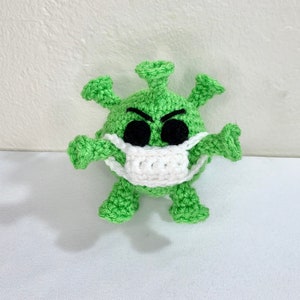 May include: Green crocheted plush toy shaped like a virus wearing a white crocheted face mask. The toy has black eyes and a frowning expression.