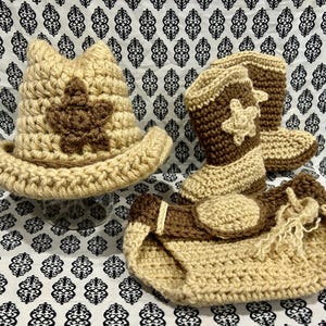 May include: A crocheted baby set in tan and brown, including a cowboy hat with a star, cowboy boots with star accents, and a diaper cover. The set is handmade with a textured stitch and is displayed on a patterned fabric.