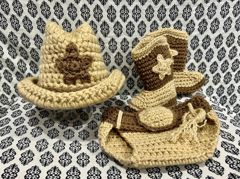May include: A crocheted baby set in tan and brown, including a cowboy hat with a star, cowboy boots with star accents, and a diaper cover. The set is handmade with a textured stitch and is displayed on a patterned fabric.