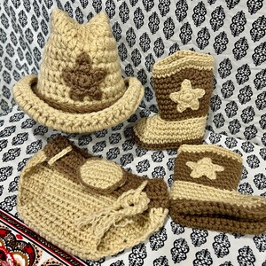 May include: A crochet baby set in tan and brown, featuring a cowboy hat with a star, two cowboy boots, and a diaper cover. The set is handmade with a star design on the hat and boots.