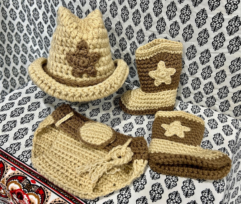 May include: A crochet baby set in tan and brown, featuring a cowboy hat with a star, two cowboy boots, and a diaper cover. The set is handmade with a star design on the hat and boots.