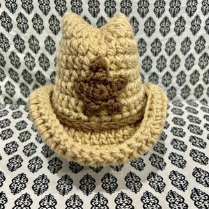May include: A crocheted cowboy hat in shades of tan and brown. The hat features a star design on the front and a wide brim. The hat is handmade and appears to be made of yarn.