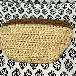 May include: A handmade, crochet pouch in a light tan color with a dark brown top. The pouch is oval-shaped and has a textured appearance due to the crochet stitches. The background is a white fabric with a black floral pattern.