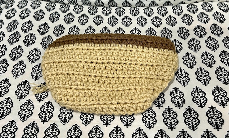 May include: A handmade, crochet pouch in a light tan color with a dark brown top. The pouch is oval-shaped and has a textured appearance due to the crochet stitches. The background is a white fabric with a black floral pattern.