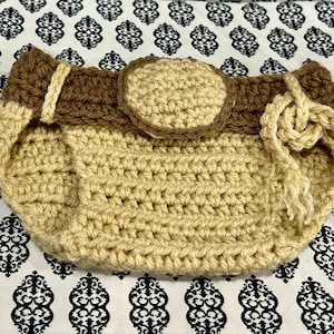 May include: Handmade crochet bag in tan and brown. The bag features a rounded shape with a brown strap and a circular accent. A knotted drawstring closure is visible on the side. The bag is set against a patterned background.