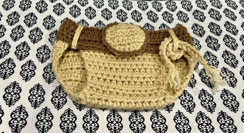 May include: Handmade crochet bag in tan and brown. The bag features a rounded shape with a brown strap and a circular accent. A knotted drawstring closure is visible on the side. The bag is set against a patterned background.