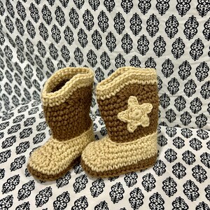 May include: A pair of handmade crochet baby boots in a cowboy boot style. The boots are primarily tan and brown, with a star-shaped embellishment on the side. The boots are displayed on a patterned white and black fabric.