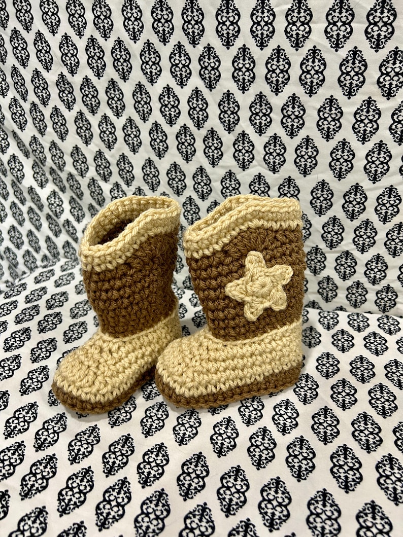 May include: A pair of handmade crochet baby boots in a cowboy boot style. The boots are primarily tan and brown, with a star-shaped embellishment on the side. The boots are displayed on a patterned white and black fabric.