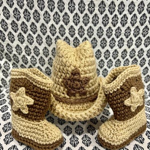 May include: A crochet baby set featuring a cowboy hat and two boots. The hat and boots are beige and brown, with a star design on the hat and boots. The set is made of yarn.
