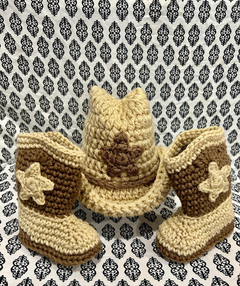 May include: A crochet baby set featuring a cowboy hat and two boots. The hat and boots are beige and brown, with a star design on the hat and boots. The set is made of yarn.