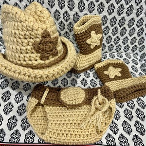 May include: A crochet cowboy hat, boots, and diaper cover set in tan and brown. The hat features a brown star, and the boots and diaper cover have star accents. The set is designed for a baby.