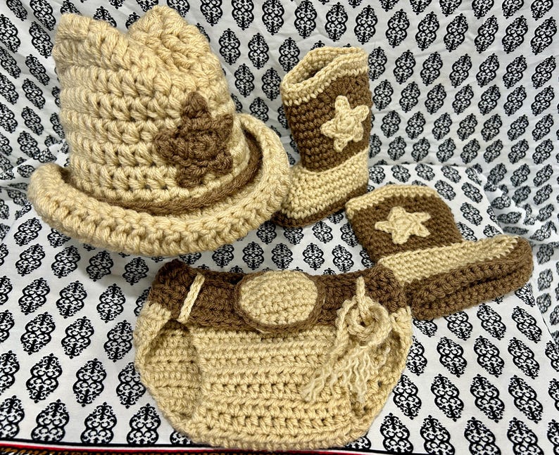 May include: A crochet cowboy hat, boots, and diaper cover set in tan and brown. The hat features a brown star, and the boots and diaper cover have star accents. The set is designed for a baby.