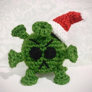 May include: Green crocheted plush toy shaped like a virus with a red and white crocheted Santa hat. The toy has a grumpy expression with black eyes and a frown.