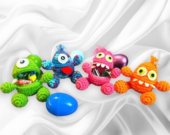 May include: Four colorful, crocheted monster-shaped candy holders. The monsters are green, blue, pink, and orange, each with a unique expression and holding candy. A blue plastic egg and a purple egg are also present.