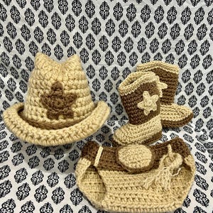 May include: A crochet baby set in tan and brown. The set includes a cowboy hat with a star, cowboy boots with star accents, and a diaper cover. The items are arranged on a patterned fabric.