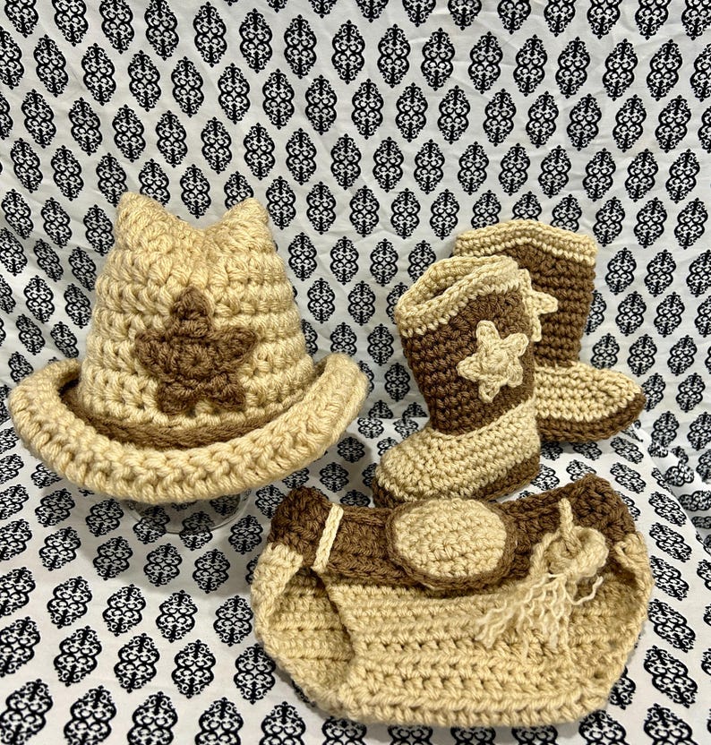 May include: A crochet baby set in tan and brown. The set includes a cowboy hat with a star, cowboy boots with star accents, and a diaper cover. The items are arranged on a patterned fabric.