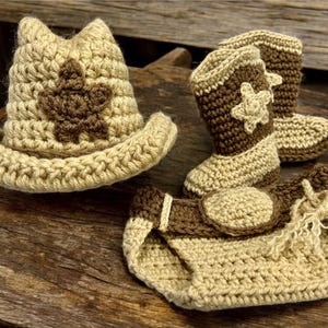 May include: A crocheted baby set in tan and brown, featuring a cowboy hat, boots, and diaper cover. The hat and boots have a star design. The set is handmade and suitable for a baby.