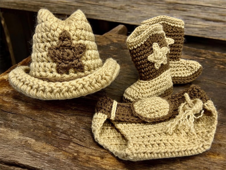 May include: A crocheted baby set in tan and brown, featuring a cowboy hat, boots, and diaper cover. The hat and boots have a star design. The set is handmade and suitable for a baby.
