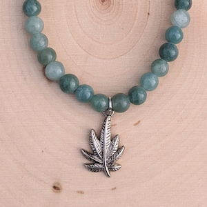 Puede incluir: Una pulsera de cuentas redondas de color verde claro. Un dije plateado con forma de hoja de cannabis cuelga de la pulsera. La pulsera se muestra sobre una superficie de madera marrón claro.