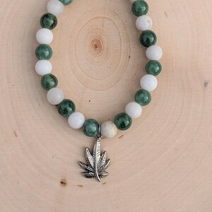 Puede incluir: Una pulsera de cuentas con cuentas redondas blancas y verde oscuro alternadas. Un dije plateado en forma de hoja de cannabis cuelga de la pulsera. La pulsera se exhibe sobre una superficie de madera clara.