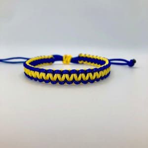 May include: A blue and yellow woven bracelet. The bracelet is made of interwoven cords in a repeating pattern of blue and yellow. The ends of the bracelet have adjustable blue cords with a black bead.