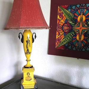 May include: A yellow table lamp with a red lampshade and a black and white design on the base. The base features a pair of swan-shaped handles and a portrait of a woman.
