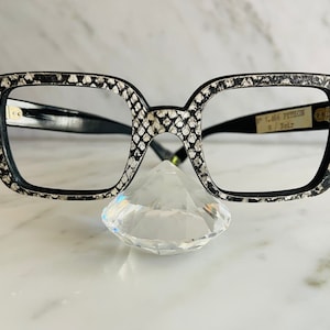 May include: Black and white square-shaped eyeglasses with a snakeskin pattern on the frame. The frame has the text "L.A.66 PYTHON W/ Noir" on the right side.